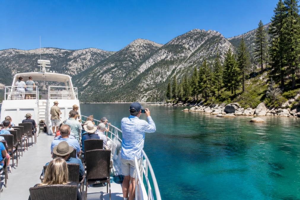 A large stable yacht cruising on crystal clear alpine lake water with Emerald Bay's granite shoreline and mountain views in the background, passengers on the open-air deck taking photos in bright midday sunlight