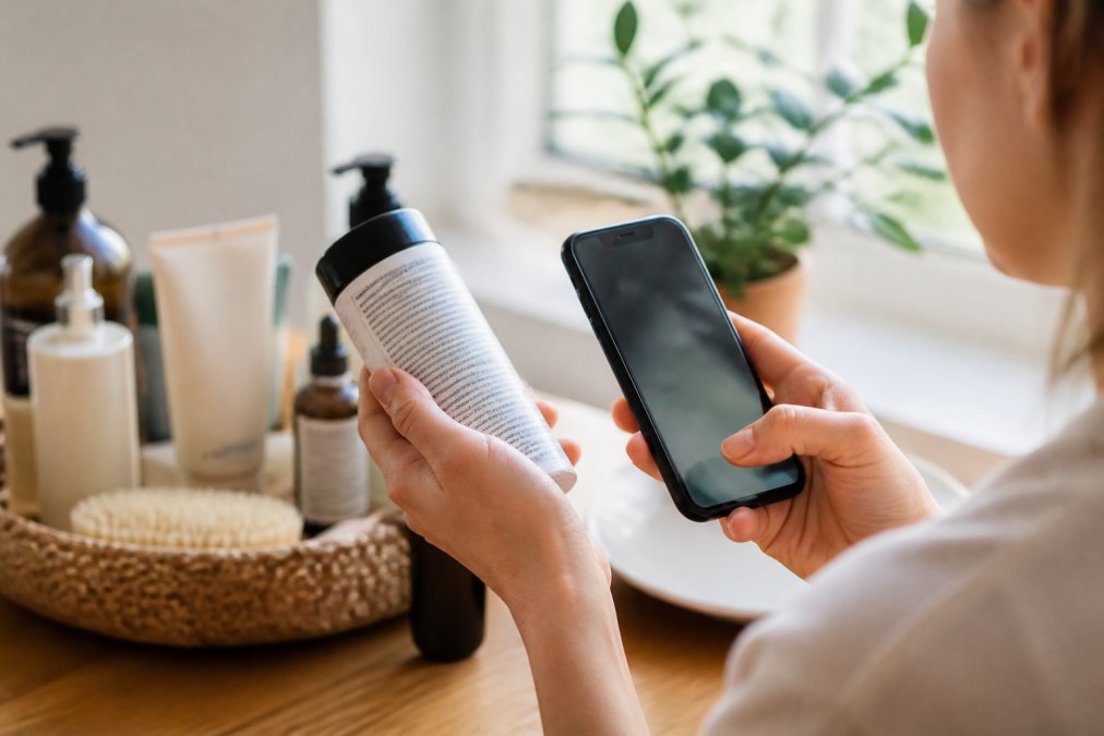Nahaufnahme einer Hand, die ein Kosmetikprodukt dreht und die Inhaltsstoffliste auf dem Etikett auf der Rückseite liest, während die andere Hand ein Smartphone mit geöffneter Scan-App hält, weiches Tageslicht