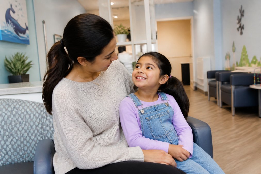 Parent and young child sitting together in a bright, calm dental waiting room with friendly, colorful decor and welcoming signage, child smiling and looking relaxed