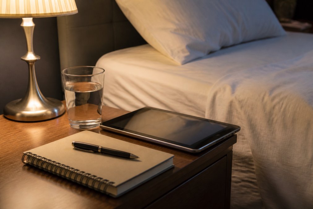 A quiet, tidy scene on a nightstand in subdued lighting; a tablet lies next to a notebook and a glass of water; in the background is a bed with clean sheets