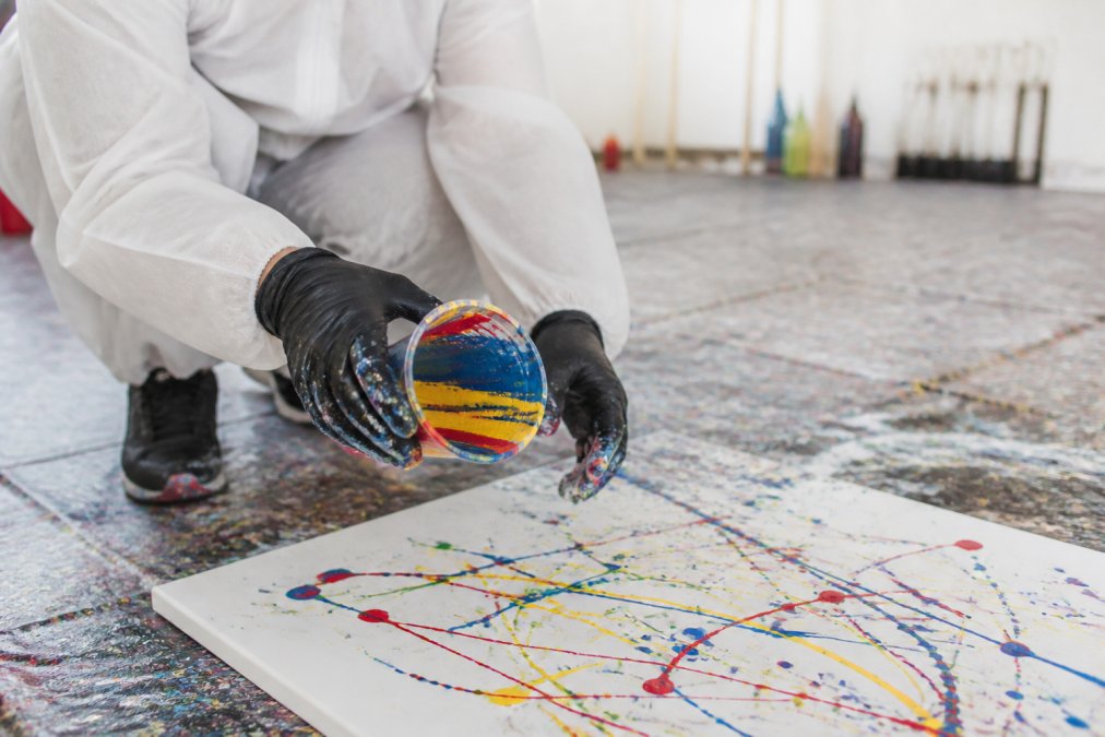Close-up of hands in a protective coverall holding a paint cup over a canvas on the floor, with colorful acrylic paint splatters visible in a bright studio