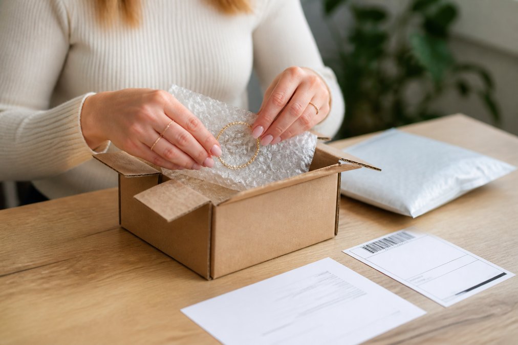 Eine Frau packt ein Schmuckstück in ein kleines Paket mit Luftpolsterfolie, auf einem hellen Holztisch liegen ein Retourenlabel und ein Lieferschein daneben