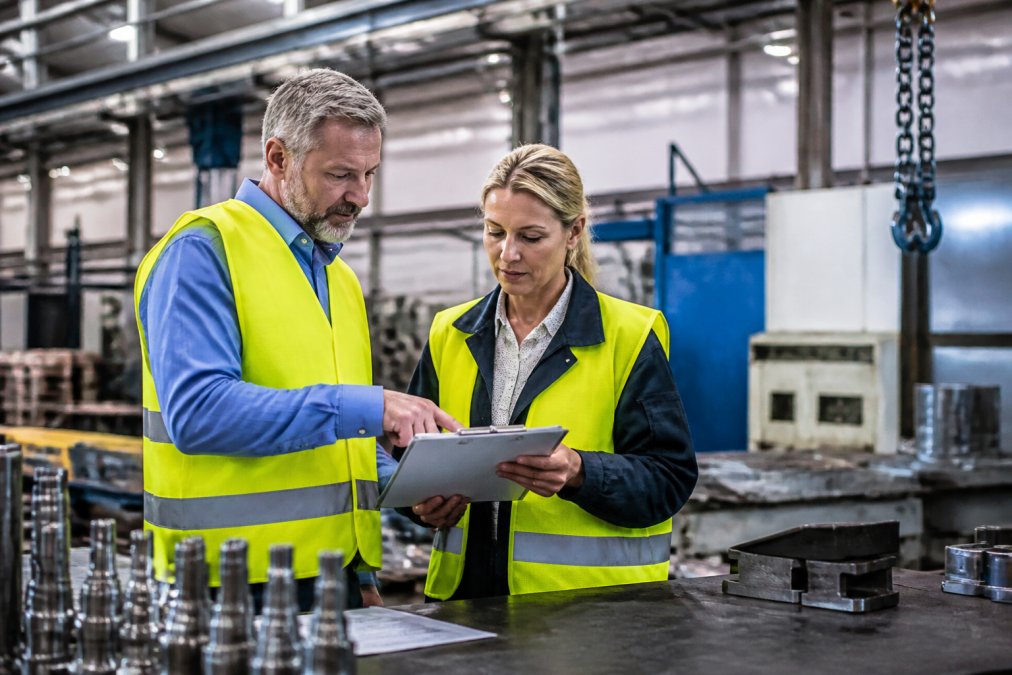 A safety officer and a production manager reviewing a checklist together at a workstation in a manufacturing facility, both wearing high-visibility safety vests, with machinery visible in the background under bright industrial lighting