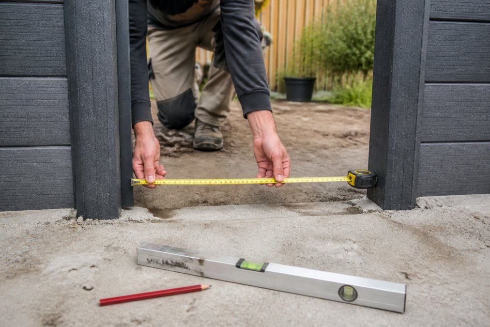 Close-up van iemand die een tuinopening opmeet met een meetlint, met waterpas en potlood zichtbaar op de grond, natuurlijk daglicht
