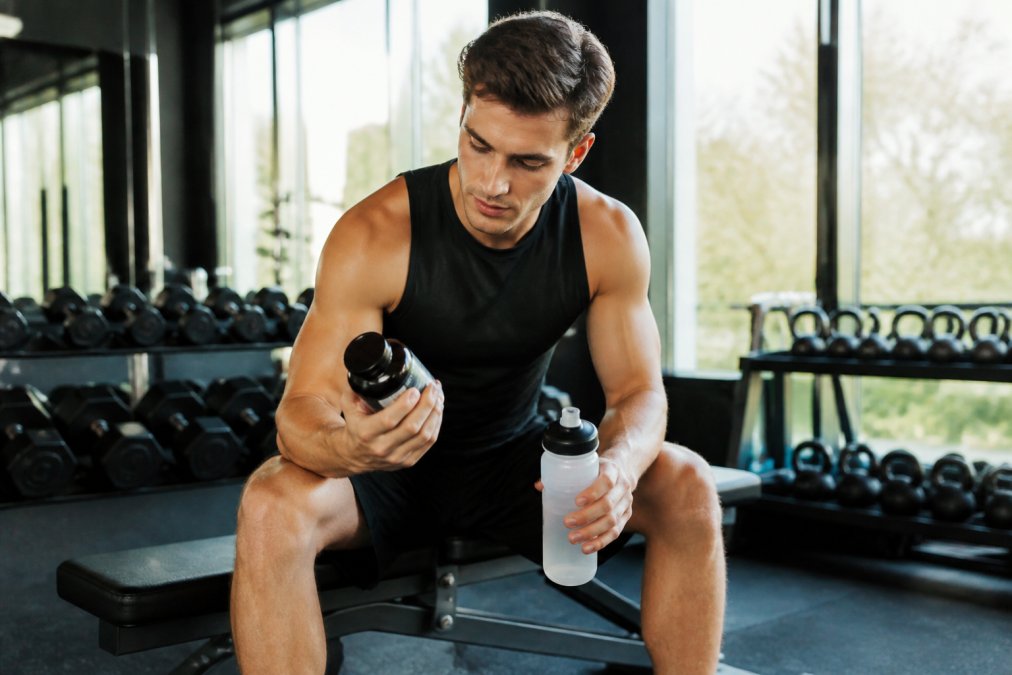 A runner in a bright modern gym sitting on a bench examining a supplement bottle label while holding a water bottle, natural daylight through large windows