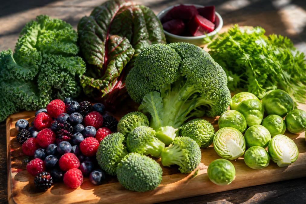Vibrant arrangement of fresh cruciferous vegetables like broccoli and Brussels sprouts, deep red berries, and dark leafy greens arranged on a wooden cutting board with natural sunlight streaming across