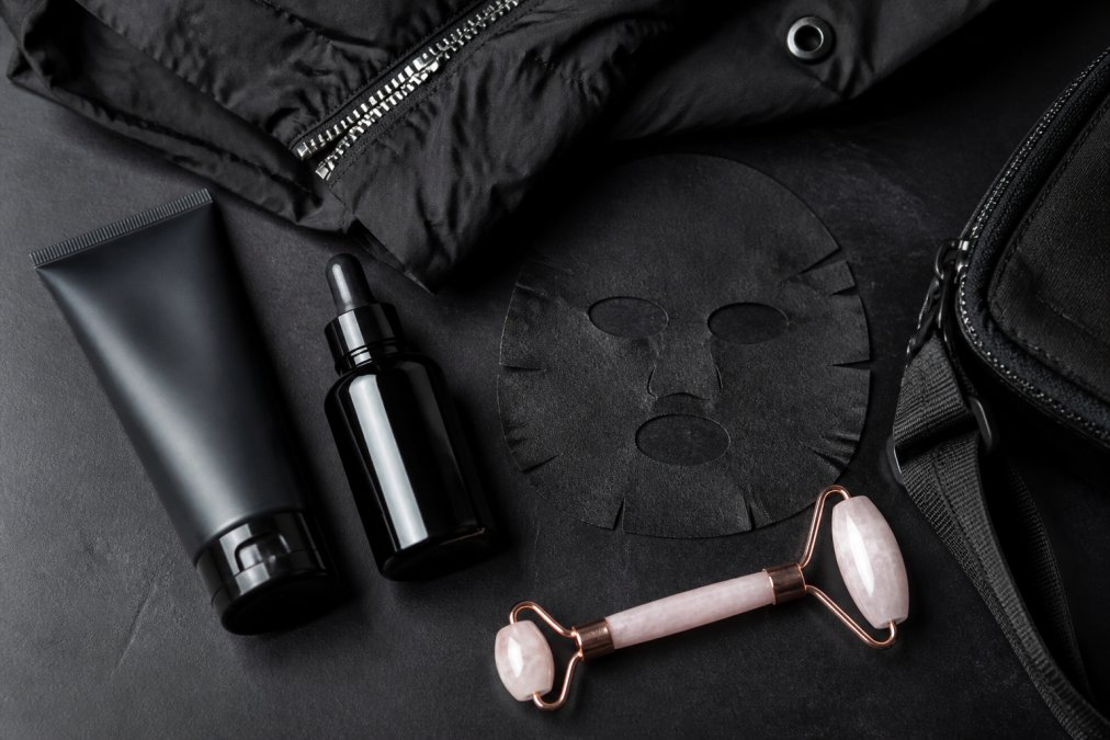 Flat-lay of skincare products including a cleanser, serum bottle, and face mask arranged on a dark matte surface alongside a folded streetwear jacket and a rose quartz roller, styled with an editorial, futuristic aesthetic under cool studio lighting