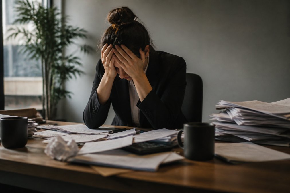 Professional in business attire sitting alone at desk with head in hands, surrounded by scattered papers and dim lighting, conveying emotional exhaustion and overwhelm in a modern office environment