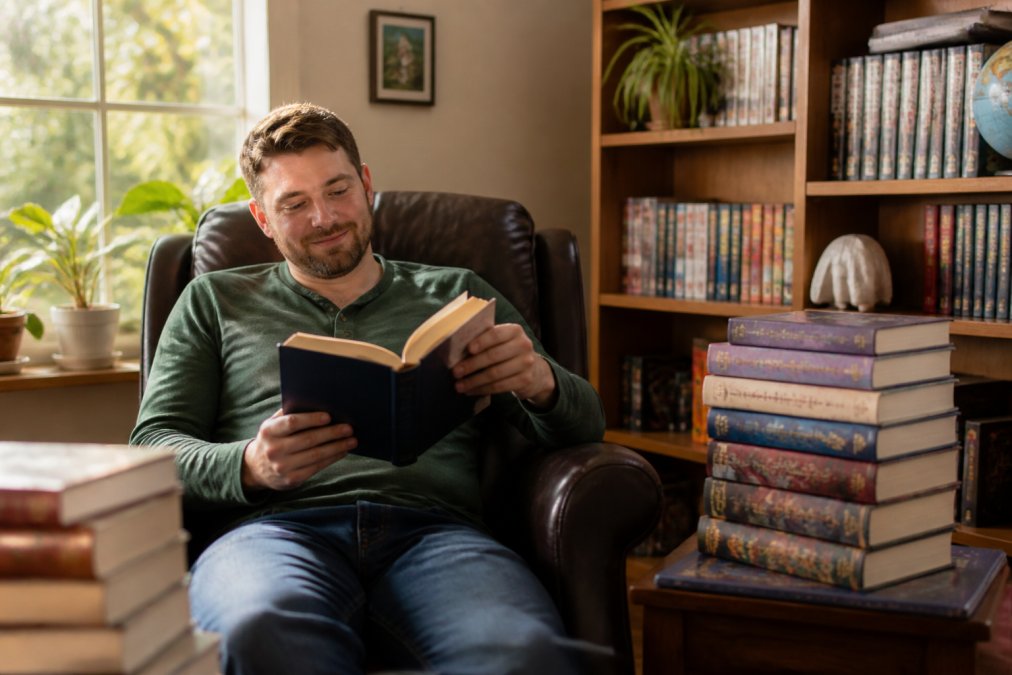 Reader sitting in a comfortable armchair with warm natural light from a nearby window, surrounded by a stack of completed science fiction book series, holding one open with visible engagement and satisfaction