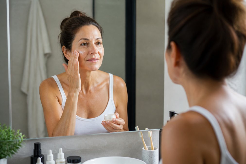 A woman in her 40s applying face cream in front of a well-lit bathroom mirror, looking calm and focused during her morning skincare routine