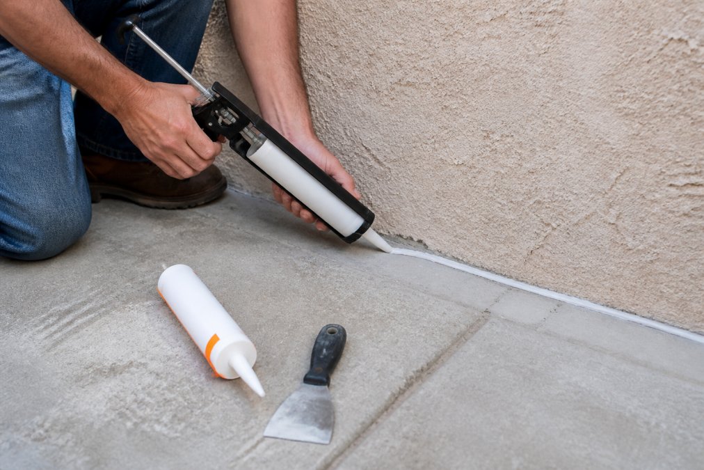 A homeowner using a caulking gun to seal a crack along the base of a stucco wall, with a tube of sealant and a putty knife nearby on the ground