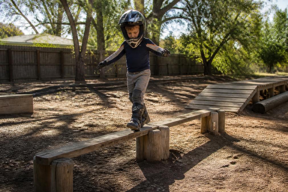 Young child balancing on a low wooden obstacle course in a backyard dirt area, wearing casual riding clothes and a helmet, with focused expression and natural afternoon sunlight casting shadows across the ground