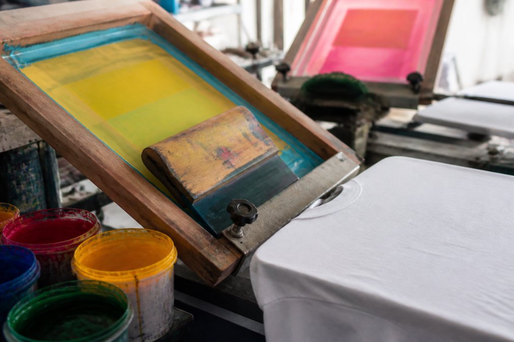 Close-up of a screen printing setup with mesh screens stretched on wooden frames, a squeegee positioned at an angle, and vibrant colored inks in containers beside a blank white t-shirt on the printing table