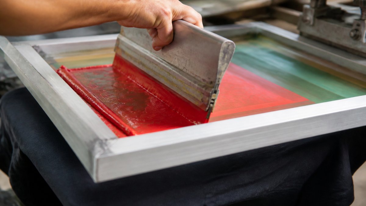 Close-up of a squeegee pressing vibrant plastisol ink through a fine mesh screen onto a dark cotton t-shirt, showing the ink flooding through the open mesh with precise color transfer and sharp image definition