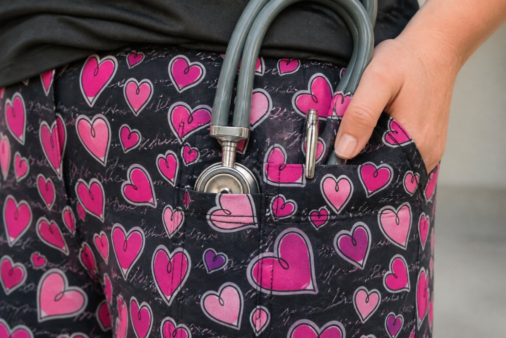 Close-up of a nurse's scrub pants pocket showing reinforced corner stitching with double-needle construction and bar tacking detail, with a stethoscope and pen resting in the pocket to demonstrate weight and daily stress