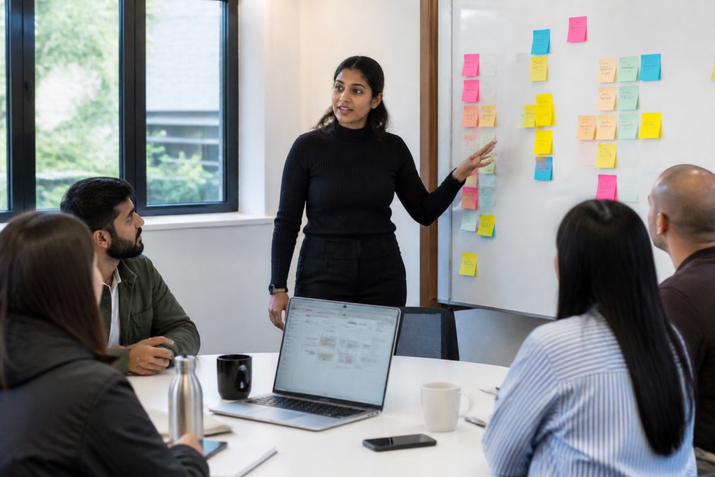 A Scrum Master standing beside a whiteboard covered in sticky notes, addressing a seated Agile team in a bright meeting room, laptop open on the table showing a sprint board