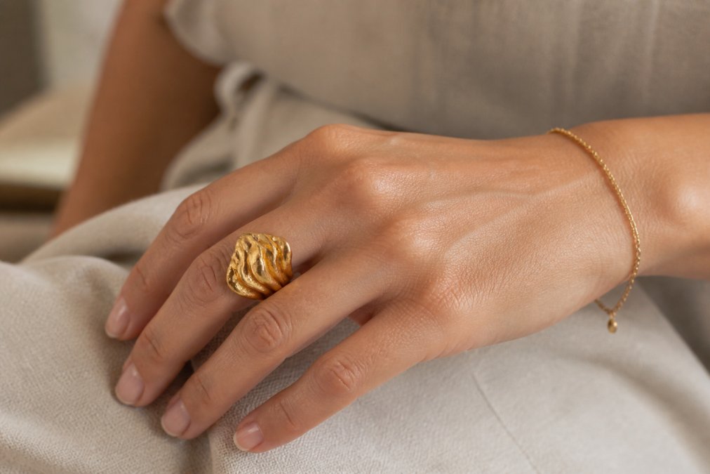Close-up of a woman's hand wearing a bold sculptural gold ring, resting on a neutral fabric surface in soft natural light, showing the ring's texture and silhouette against a simple outfit