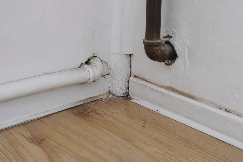 Close-up of apartment wall corner showing visible gaps, cracks, and crevices near baseboards where pipes penetrate the wall, with natural lighting highlighting problem areas