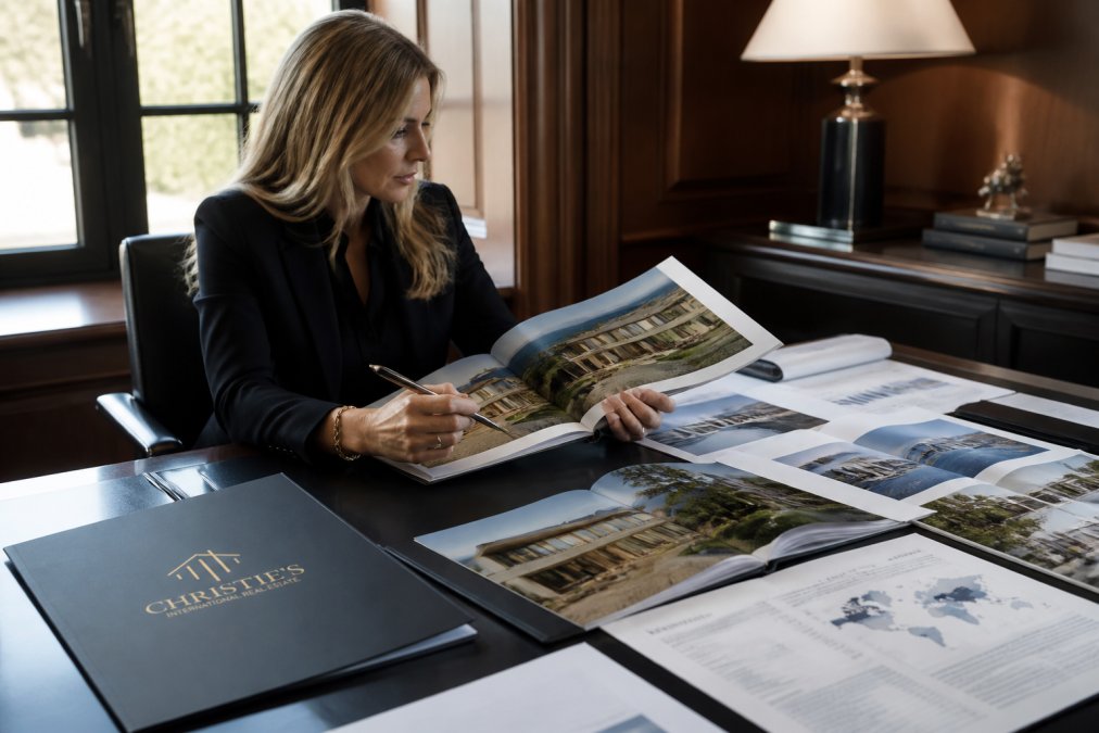 Real estate professional in upscale office reviewing luxury property marketing materials on multiple screens, with architectural renderings and market analysis documents visible on desk