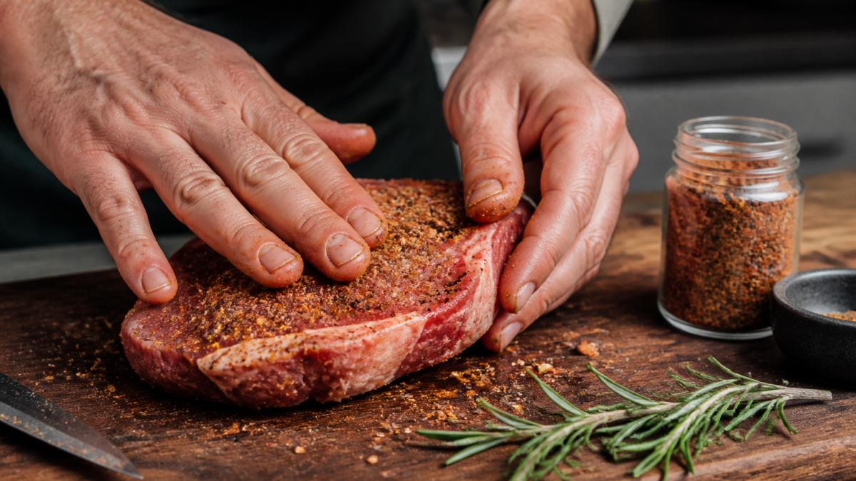 Close-up of hands generously rubbing seasoning blend onto a raw steak on a wooden cutting board, with a jar of seasoning and fresh rosemary sprigs nearby, natural kitchen lighting