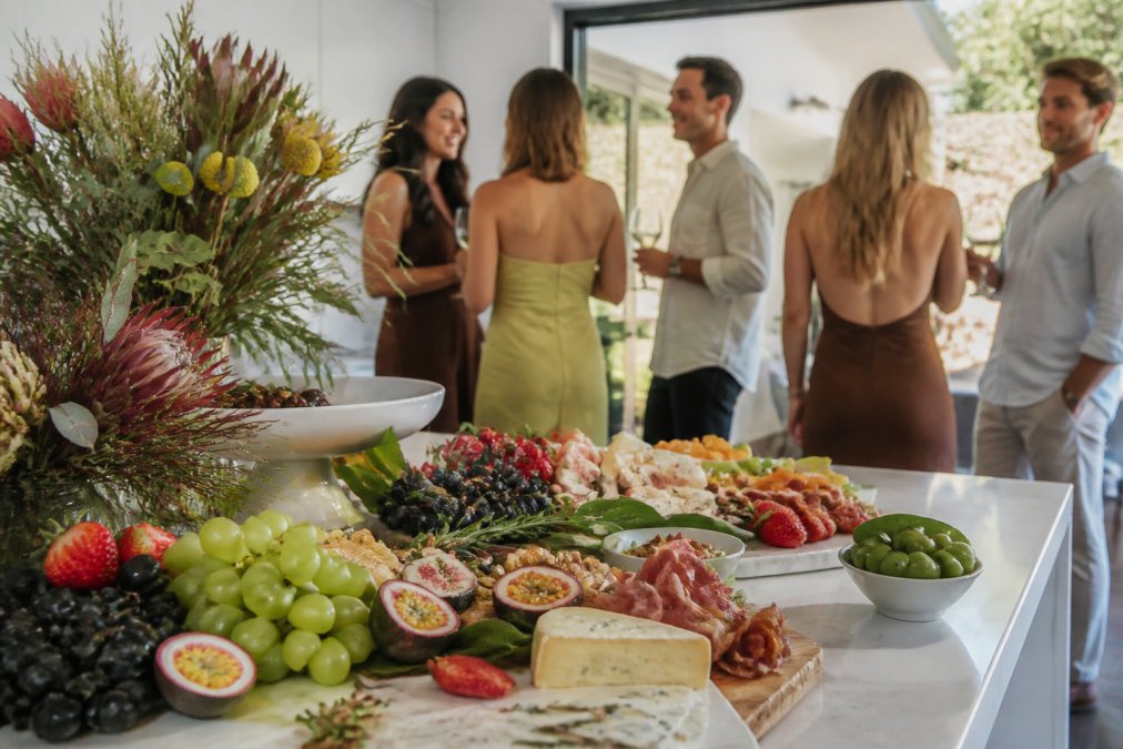 An elegant grazing table set for an engagement party in a Sydney home, with fresh seasonal fruit, cured meats, cheeses, and native Australian flowers as a centrepiece, guests mingling in the background