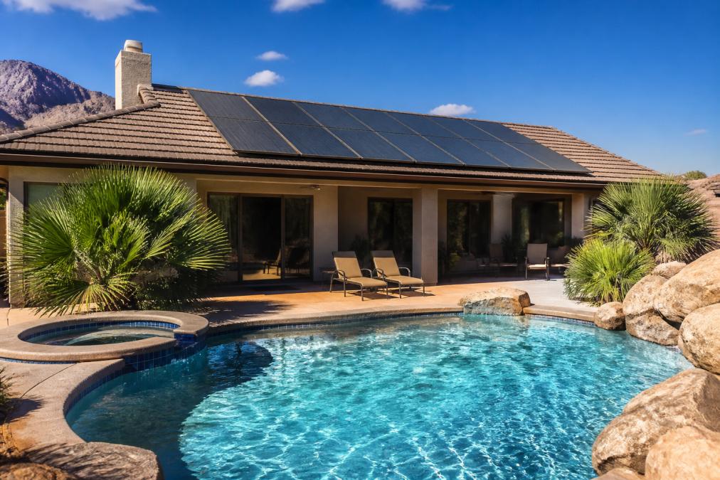 Solar thermal panels installed on a residential rooftop overlooking a backyard swimming pool, with clear blue sky and sunlight reflecting off the panels and water