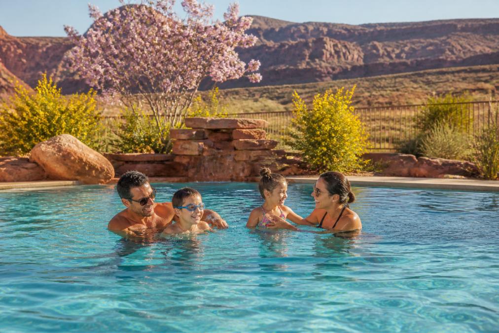A family enjoying a heated swimming pool in early spring with desert landscape, warming sunlight, and blooming native plants visible in the background