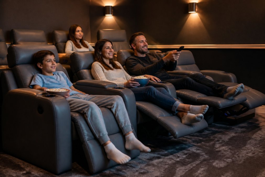 A family relaxing in a modern home theater room with power recliners in gray upholstery, showing someone using a remote control with cup holders and warm ambient lighting visible in the background
