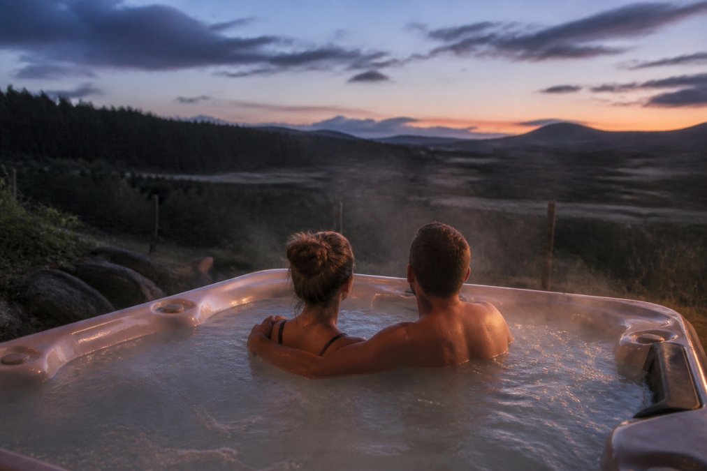 Couple relaxing together in a private outdoor hot tub at dusk, steam rising from the water, surrounded by rolling countryside and woodland, soft twilight lighting casting long shadows