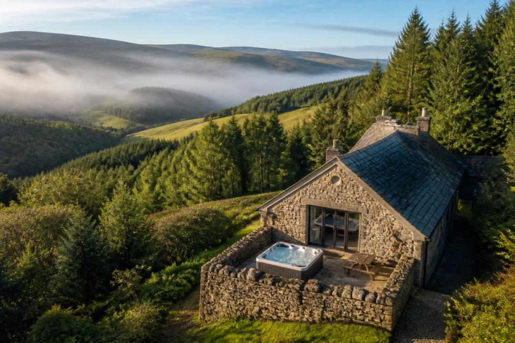Aerial view of a secluded stone cottage nestled in rolling green hills with woodland surrounding it, a private hot tub visible on the stone terrace, morning mist in the valleys below