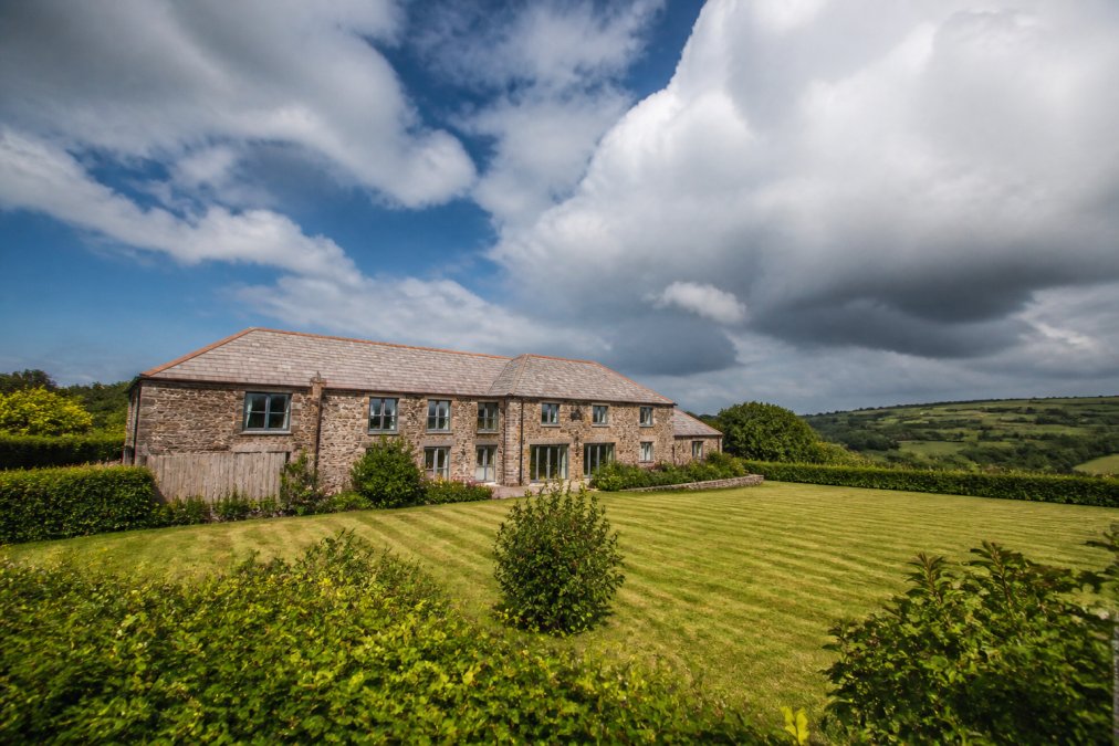A beautifully converted stone barn set in rolling South Devon countryside with manicured gardens, fields beyond, and dramatic sky, showing the property's rural isolation and architectural character
