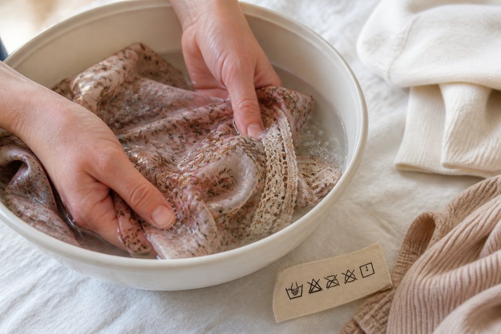 Nahaufnahme von Händen, die ein feines Vintage-Kleidungsstück in einer Schüssel mit klarem Wasser vorsichtig von Hand waschen, daneben ein Pflegeetikett mit Wäschesymbolen, heller Hintergrund