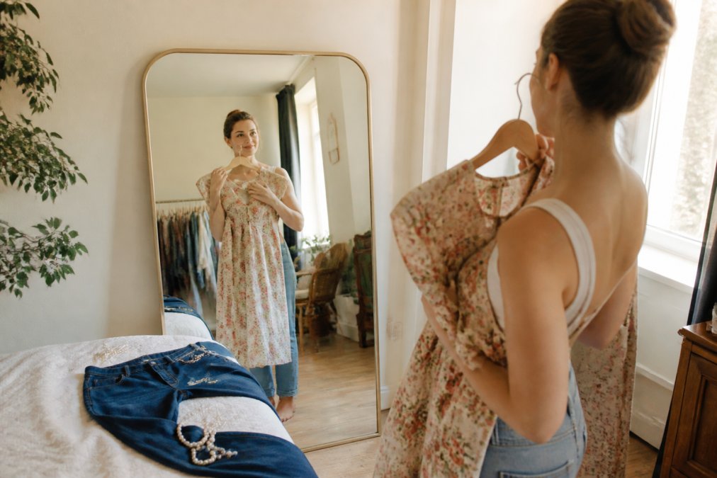 Frau kombiniert Second Hand Mode mit einem Vintage-Kleid, Jeans und Perlenkette vor einem Spiegel.