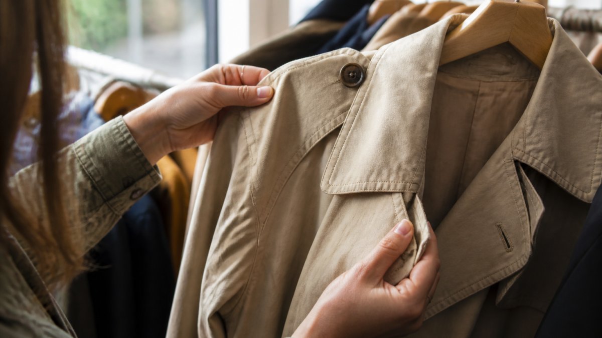 Nahaufnahme von Händen, die ein Second-Hand-Kleidungsstück unter natürlichem Licht genau untersuchen - Fokus auf Nähte, Knöpfe, Stoff-Oberfläche und mögliche Verschleißstellen