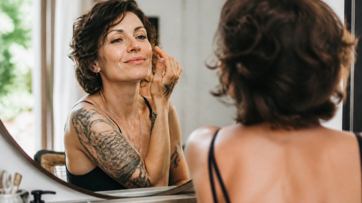 Woman in her 40s smiling confidently at herself in a mirror with natural morning light, relaxed and comfortable in her own skin, wearing minimal makeup