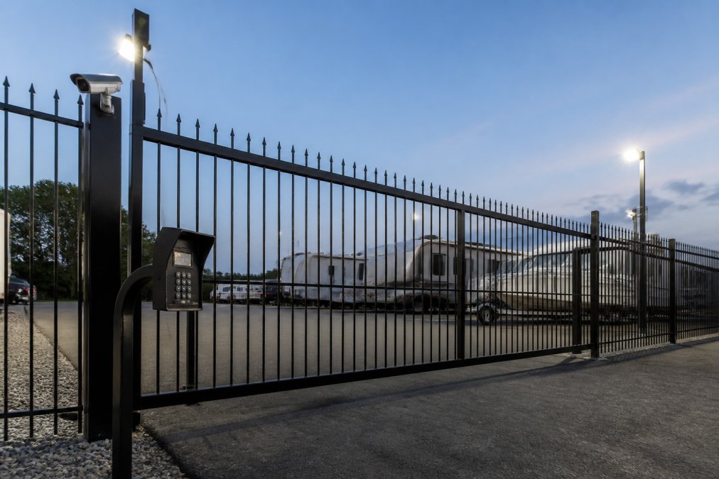 Wide shot of a gated storage facility entrance with security lighting, keypad entry system, and 24/7 surveillance camera mounted on the fence