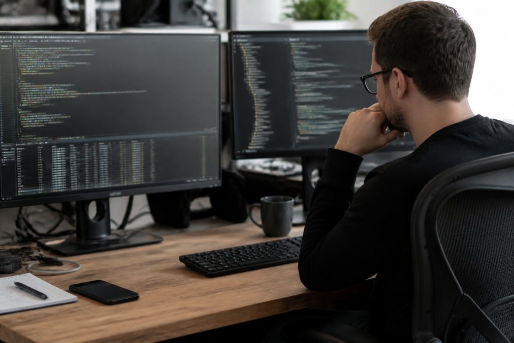 Security professional at desk with multiple monitors displaying code and network logs, focused expression as they review browser developer tools and security audit results