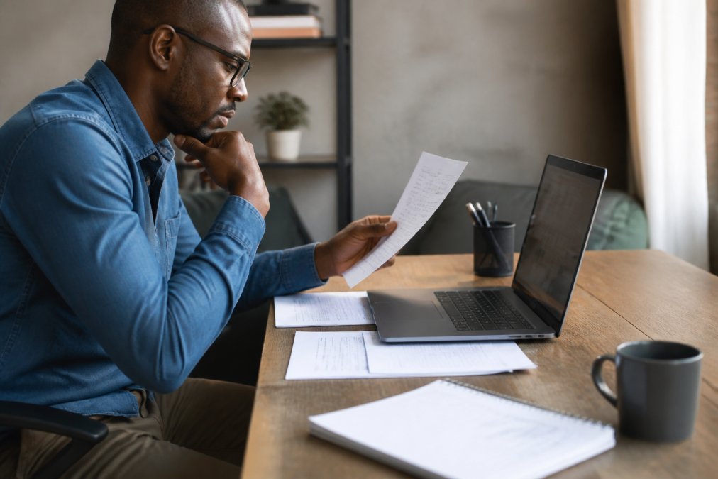 Professional sitting at desk reviewing computer screen with documents and notebook, focused expression while checking personal information
