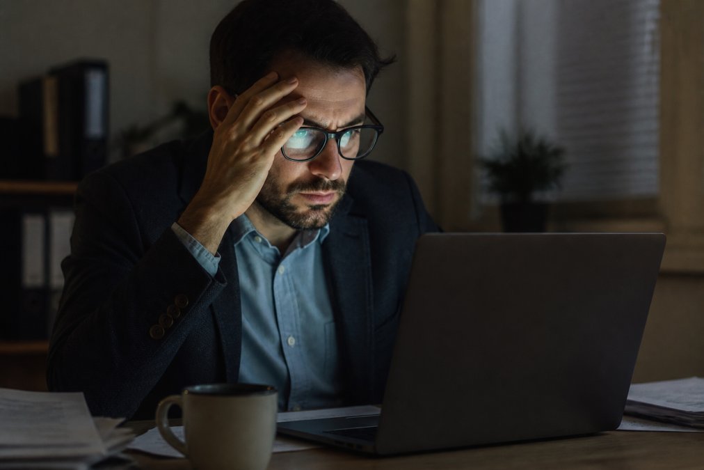 A professional looking concerned while reviewing a security warning on a laptop screen, face illuminated by the screen glow in a dimly lit office with papers and a coffee cup on the desk