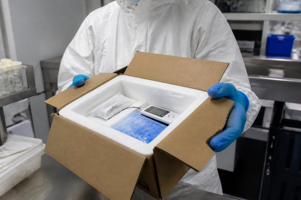 A gloved laboratory researcher in a cleanroom setting holding an opened shipping box lined with thick foam insulation and a temperature data logger, with cold packs visible inside under bright overhead lighting