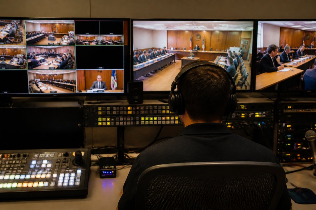 Control room operator monitoring multiple screens displaying live video feeds, encoding metrics, and streaming statistics during a government meeting broadcast, with professional lighting and technical equipment visible