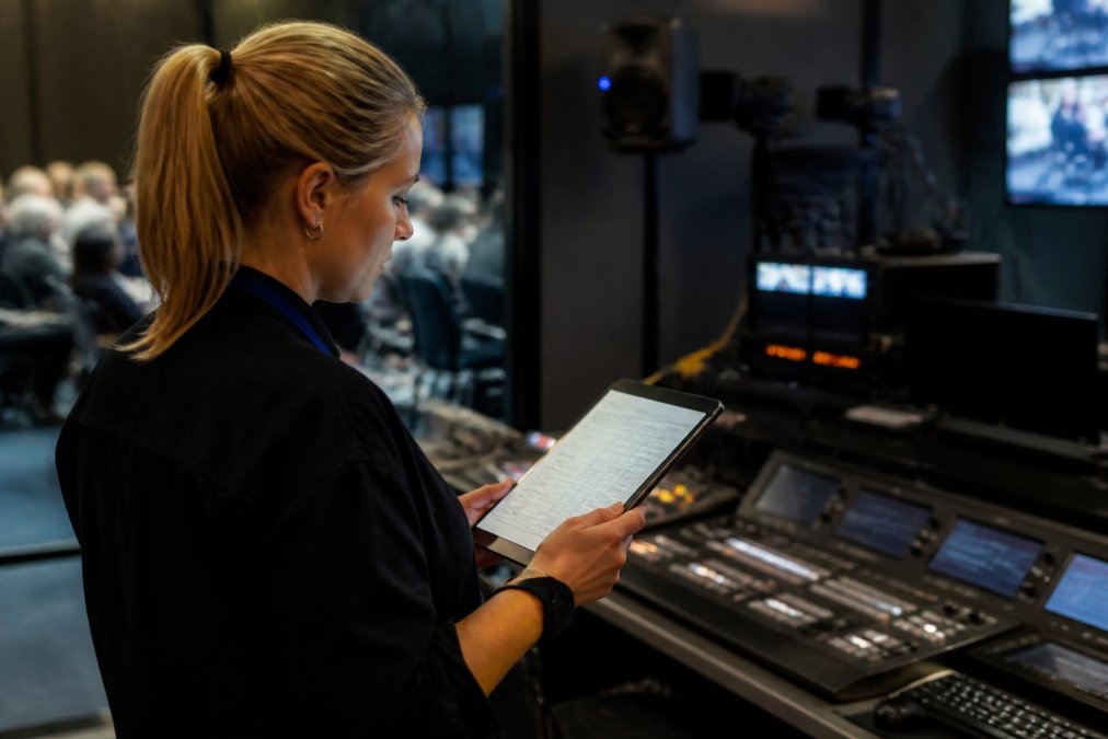 Corporate event coordinator reviewing attendee access credentials and participant list on a tablet before a town hall broadcast in a professional control room