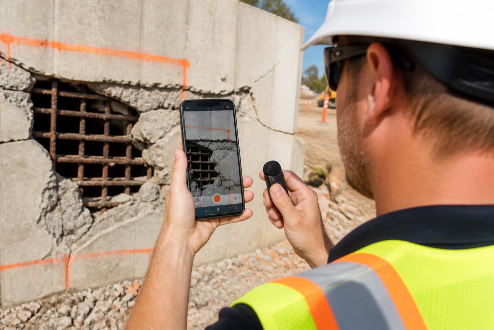Field inspector in reflective safety vest holding an Android smartphone while documenting a construction site defect, with geotagged photo visible on the screen and voice notes being added in bright daylight