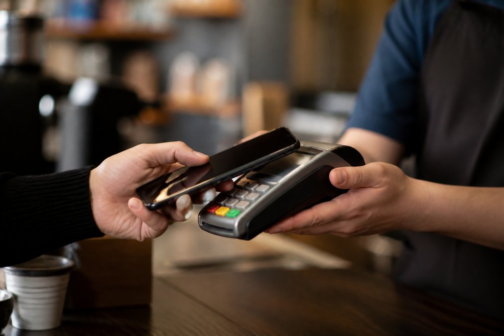 Person's hand holding smartphone near a payment terminal, showing mobile wallet contactless payment in progress at a retail counter with warm indoor lighting
