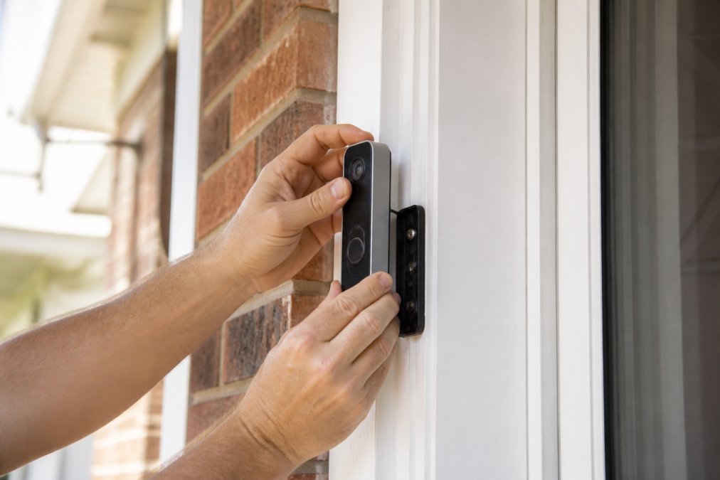 A homeowner's hands installing a battery-powered doorbell camera on an entryway door frame, showing the mounting bracket being secured with screws and the camera clicking into place, with natural daylight illuminating the installation