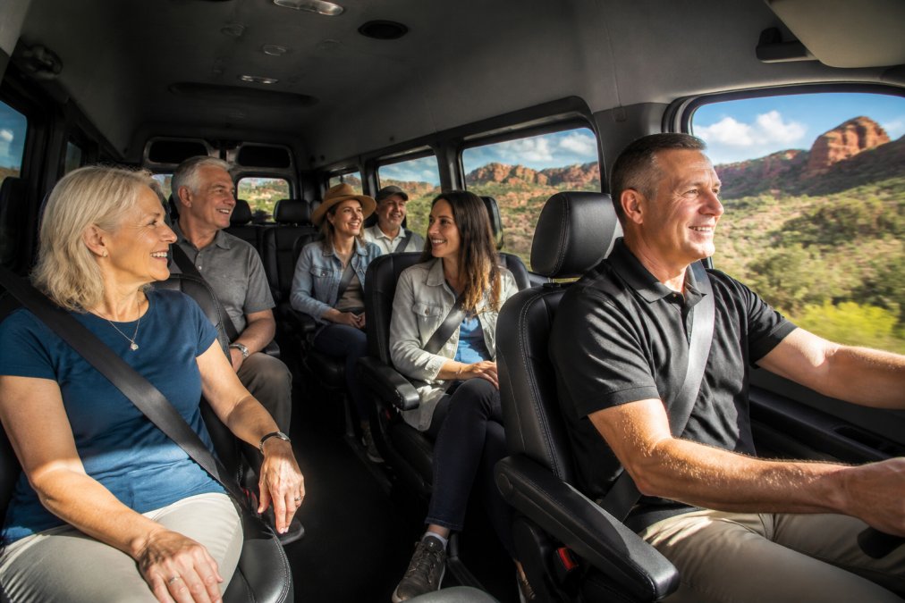 Small group of travelers in a comfortable Ford Transit vehicle with flexible seating, relaxed and smiling, driving through red rock desert landscape with professional guide visible in driver's seat