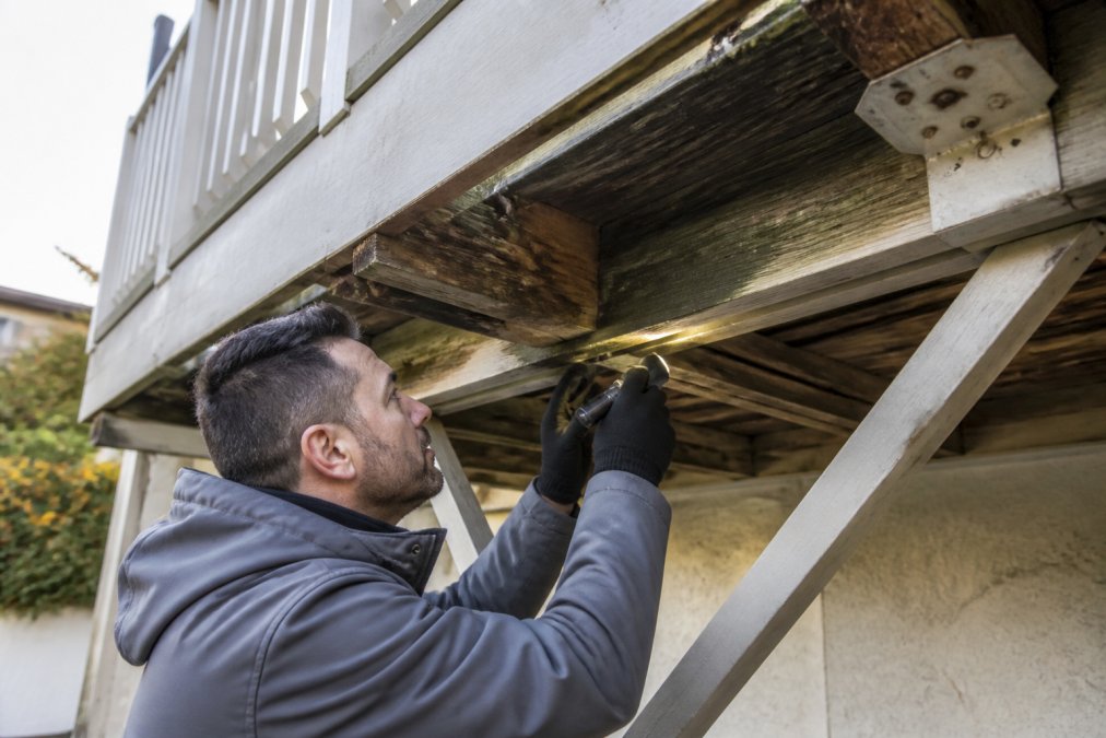 Professional building inspector examining wooden balcony structure from underneath, using flashlight and inspection tools to check for decay and fastening failures on a residential building
