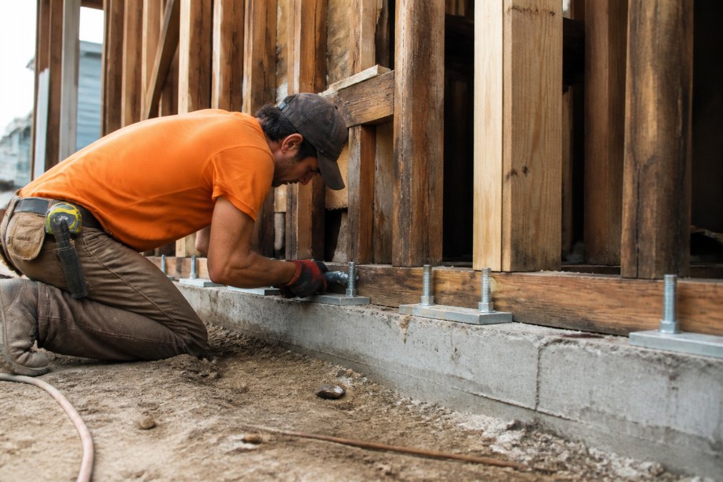 Professional contractor installing anchor bolts and foundation connections on concrete foundation of older home during seismic retrofit work, with exposed framing and tools visible