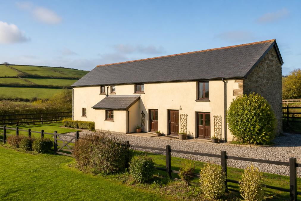 Stone barn exterior with cream rendering, dark wooden doors and windows, surrounded by manicured gardens with secure fencing visible, countryside rolling hills in soft afternoon light