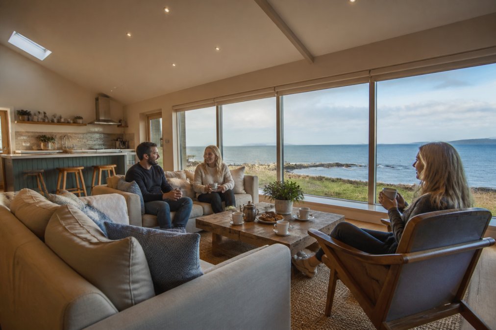 A bright, spacious self-catering lodge living area with a small group relaxing on sofas, a kitchenette visible in the background, and large windows framing a view of the Isle of Skye coastline